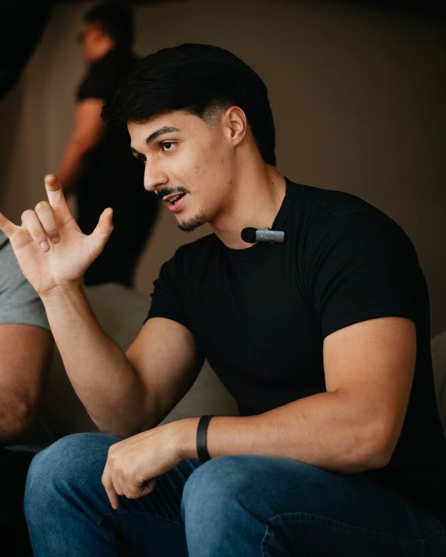 Tony Alves sentado, de camiseta preta e microfone de lapela, fazendo um sinal em Libras enquanto fala