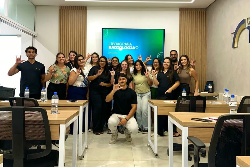 Turma de profissionais de saúde, vários de uniforme, fazendo um sinal de Libras junto com o Tony numa sala de treinamento
